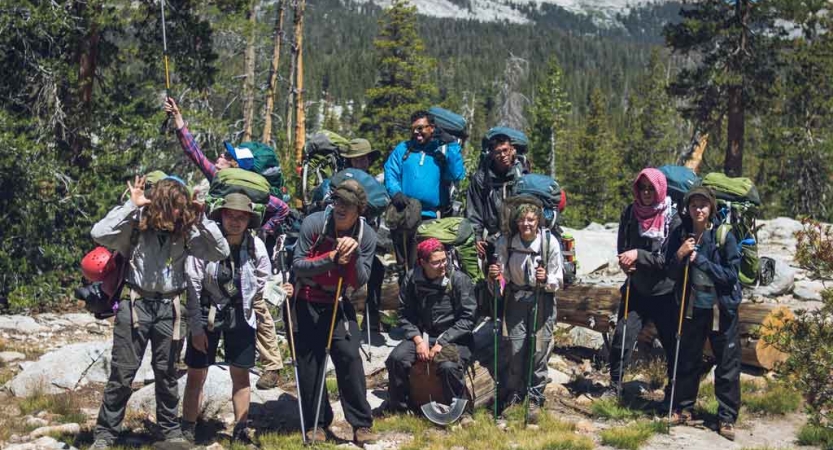 A group of people wearing backpacks pose for a group photo in a green wooded area. 